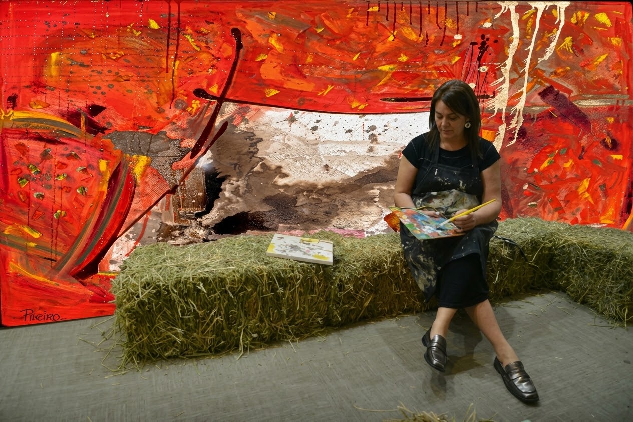 Gabriela Rieiro con un cuaderno de bocetos, sentada frente a una de sus obras de gran formato